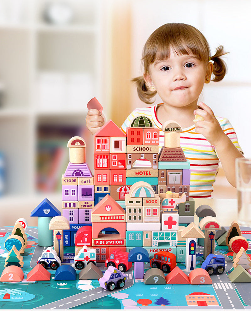 A young child plays with a colorful set of InvenToy Large Montessori Wooden Building Blocks and vehicle toys, including buildings labeled as "museum", "school", and "cafe", arranged on a play mat with roads and numbers.