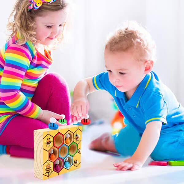 Two young children, a girl and a boy, play together with a colorful InvenToy Montessori Bee Box on the floor. The girl wears a striped dress while the boy is in a blue shirt.