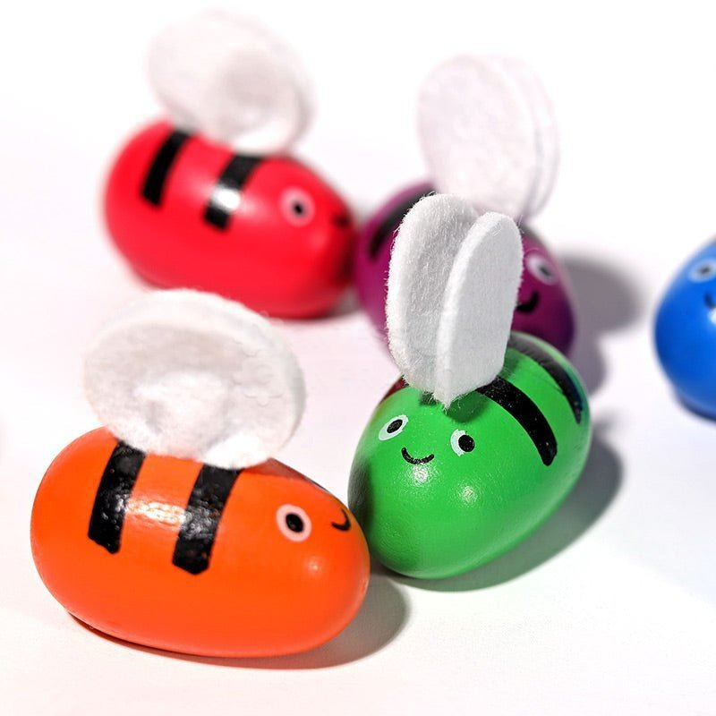 Colorful InvenToy Montessori Bee Boxes with smiling faces and fluffy white antennae are arranged on a white background, featuring bright red, green, purple, and blue colors.
