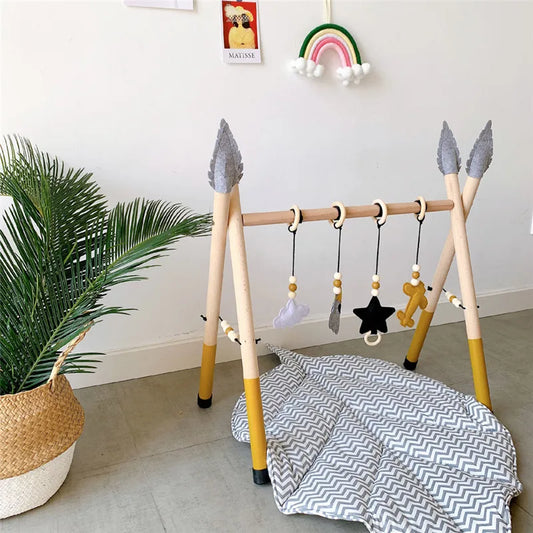A stylish InvenToy wooden exercise machine stands over a zigzag-patterned mat, featuring dangling wooden toys and plush shapes, in a bright room with a potted plant and rainbow wall decor.