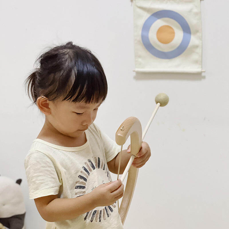 A young child with black hair and bangs is focused on playing with the InvenToy Montessori Bow and Arrow Set, enhancing their hand-eye coordination. Behind the child, there is a wall hanging with a target design consisting of concentric circles in blue, white, and yellow. The background is plain and off-white.