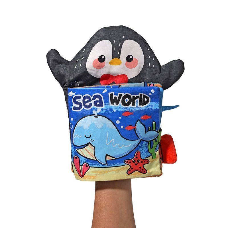 A Montessori Baby Cloth Book hand puppet shaped like a cartoonish penguin wearing a bow tie, holding a colorful "sea world" book with illustrations of marine animals, designed by InvenToy.