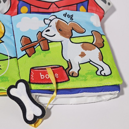 A colorful InvenToy Montessori Baby Cloth Book open to a page featuring a cartoon illustration of a happy dog with brown spots, next to a red bowl labeled "bone" and a bone-shaped bookmark.