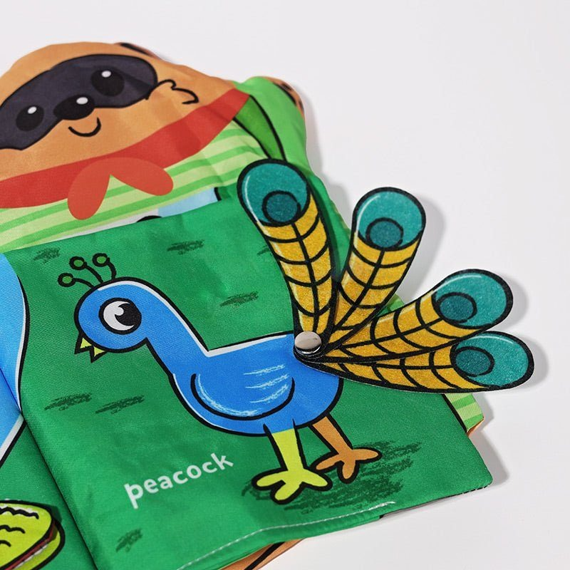 A colorful InvenToy Montessori baby cloth book opened to a page featuring a cartoon illustration of a blue peacock with expanded tail feathers, alongside a sloth peeking from an earlier page.