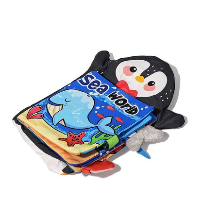A colorful children's Montessori Baby Cloth Book with a plush penguin cover depicting a cartoon penguin and an "Inventoys sea world" theme, laid on a bright background. The book shows marine animals.
