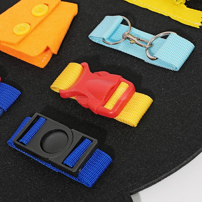 Close-up view of various colorful InvenToy Montessori Dino Busy Board baby toys including a red buckle, blue strap, yellow zipper, and orange button on a black background.