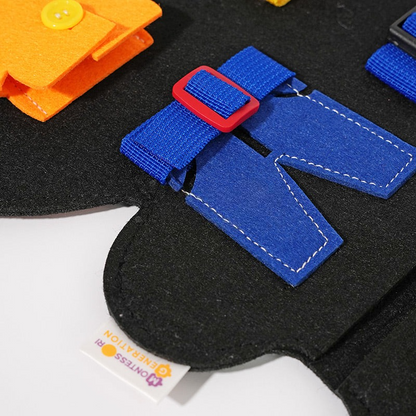 Close-up of a child's InvenToy Montessori Dino Busy Board in black with textured elements including a blue buckle, orange snap, and a clothing fastener made of blue fabric.