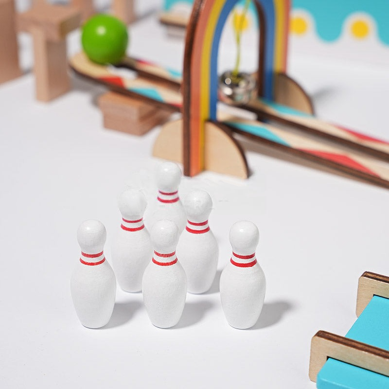 Small white bowling pins with red stripes set up in a triangle formation on a table surrounded by colorful InvenToy Montessori Domino Run and a rainbow arch.