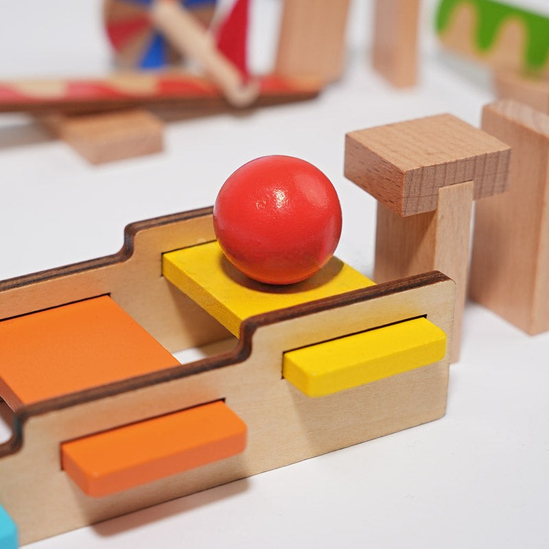 A close-up of a colorful InvenToy Montessori Domino Run wooden puzzle toy with a red ball centered on a shaped block surrounded by various other geometric shapes and toy parts.