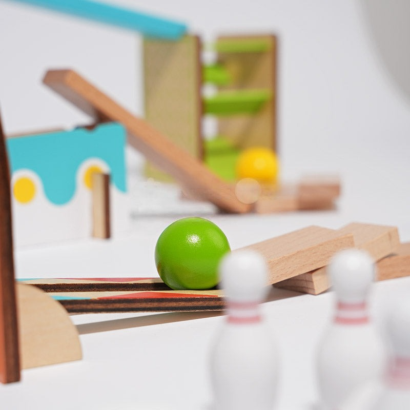A close-up image of a green ball on a wooden track part of an InvenToy Montessori Domino Run set with other elements like ramps, a yellow ball, and bowling pins blurred in the background