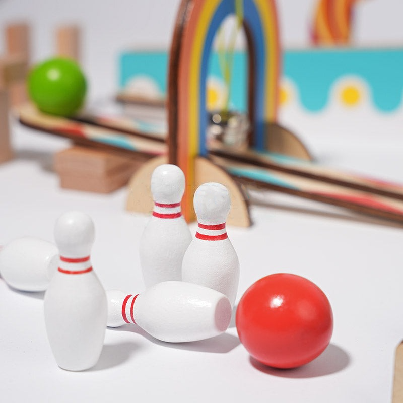 Miniature bowling pins and a red ball on a table with a colorful toy rainbow and other InvenToy Montessori Domino Run toys in the background.