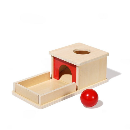A InvenToy Montessori Object Permanence Box featuring a house-shaped box with a red entrance and a round hole on top, accompanied by a single red ball on a white background.