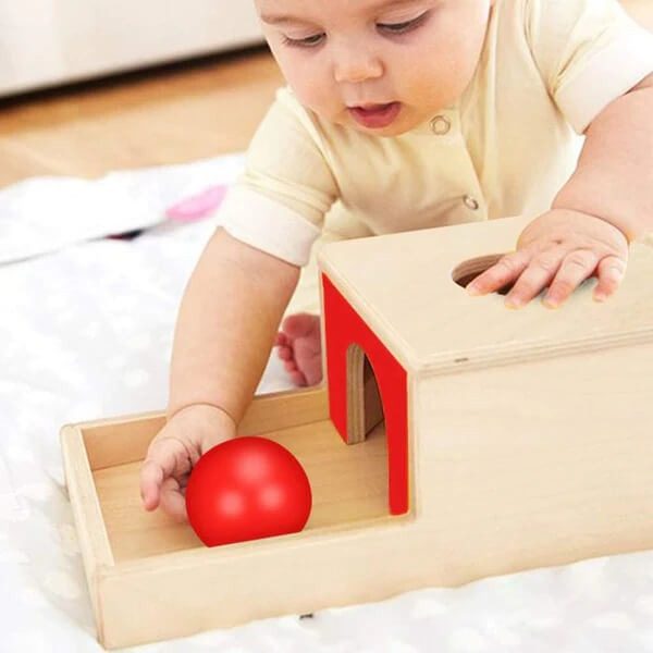 A baby engages with an InvenToy Montessori Object Permanence Box, placing a red ball through a hole in a small house-shaped structure on the floor.