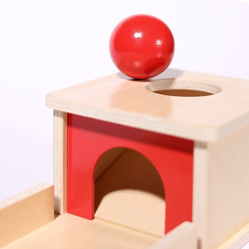 A red ball poised at the edge of a hole on a wooden block puzzle with a red and natural wood color scheme, designed as a InvenToy Montessori Object Permanence Box baby toy.