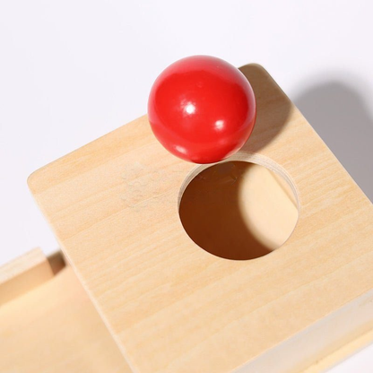 A bright red InvenToy Montessori Object Permanence Box positioned on top of a wooden board, which has a circular hole slightly smaller than the ball. The shadow of the ball is visible on the white surface.