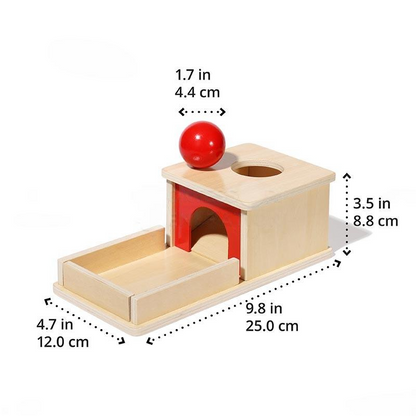 A wooden Montessori InvenToy Object Permanence Box consisting of a cube with a hole on top and a red ball above it, next to a ramp leading from the cube. Dimensions are labeled in inches and centimeters.