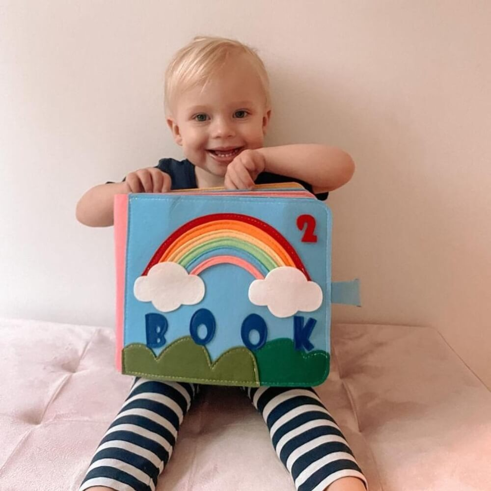 A joyful toddler with blond hair smiles while leaning on a blue Montessori Story Book labeled "InvenToy" featuring a rainbow and the word "book" on the cover. The child is wearing a dark