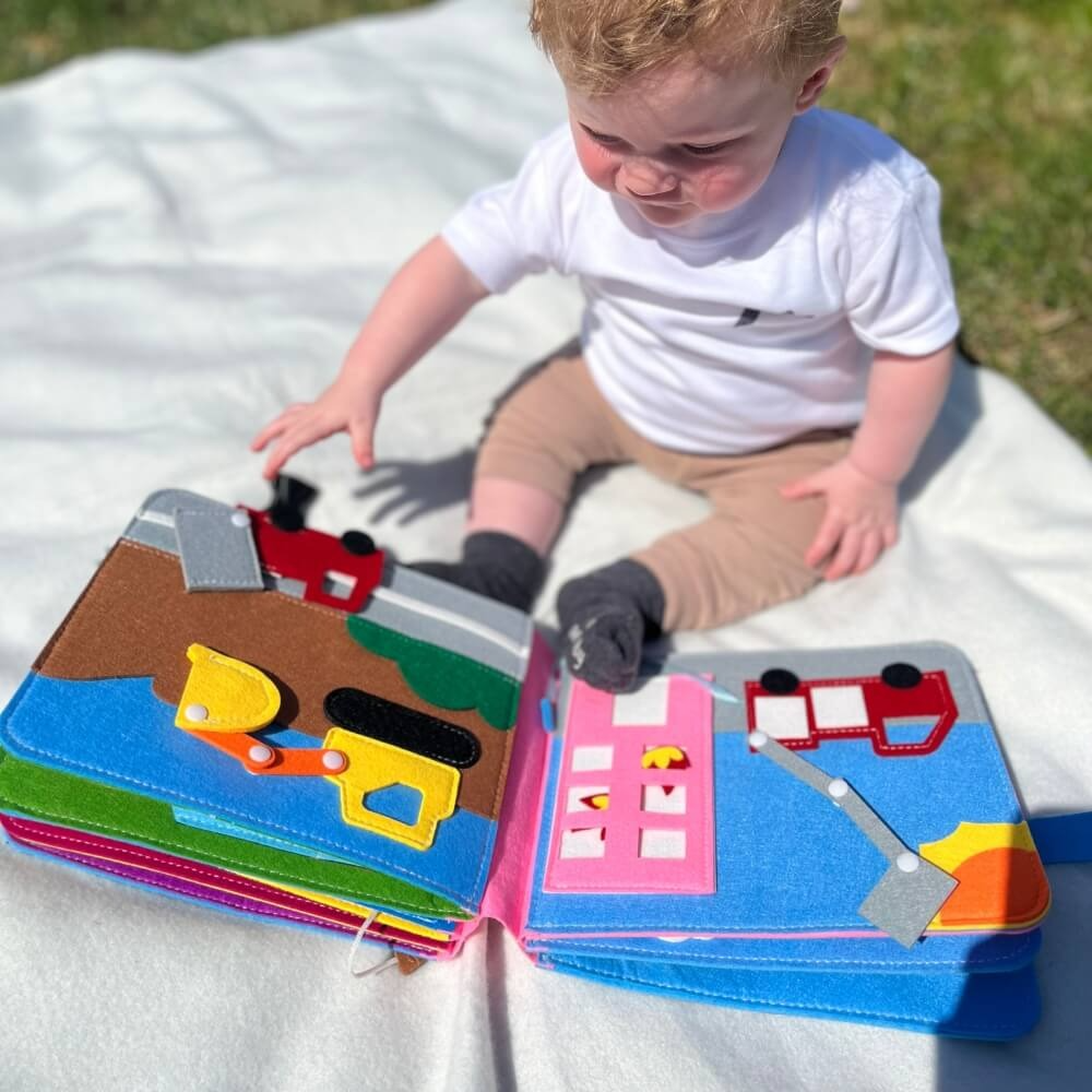 A toddler sits on grass engaging with a colorful InvenToy Montessori Story Book, focusing intently on attaching shaped pieces to matching outlines. Sunlight enhances the vivid colors of the book and the child.
