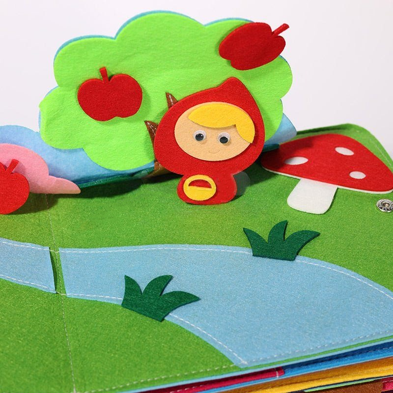Colorful children's Montessori Story Book page featuring an InvenToy scene with a small character in a red hood, apples, a mushroom, and a tree on a background of a green meadow and blue sky.