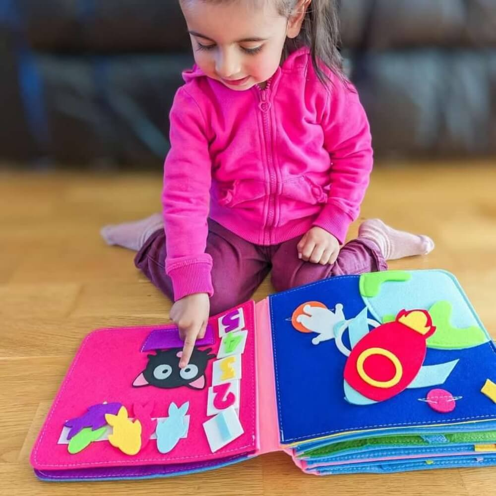 A young girl in a pink jacket is sitting on a wooden floor, interacting with a colorful, open InvenToy Montessori Story Book featuring animal shapes.