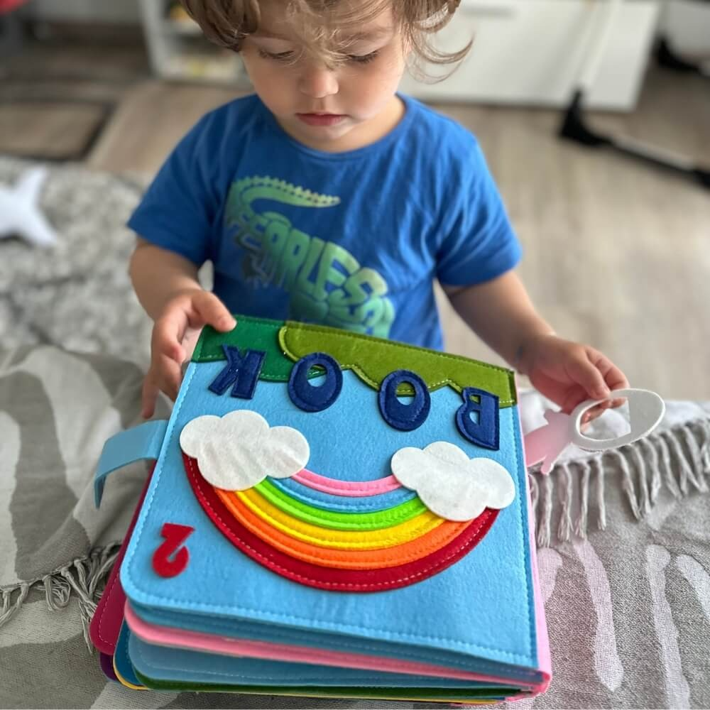 A toddler in a blue shirt reads a colorful felt InvenToy Montessori Story Book featuring a rainbow and clouds on the cover. The child is seated on a gray surface, focused on the book.