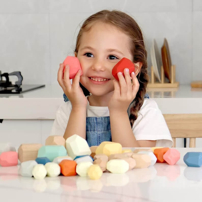 A young girl with braided hair smiles while playing with colorful InvenToy Montessori wooden stones in a kitchen, holding two red stones up to her cheeks.