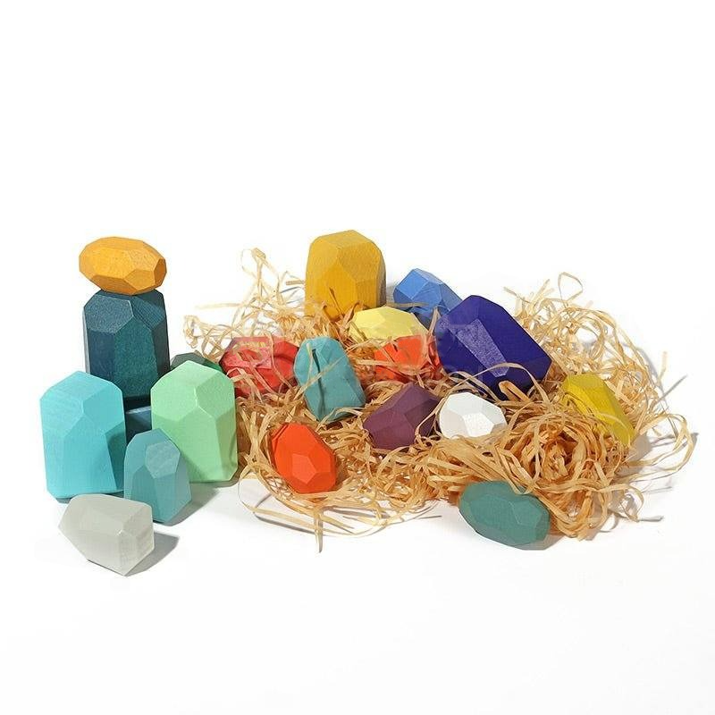 A collection of colorful, geometric InvenToy Montessori Wooden Stones (21 Pieces) scattered on straw, showcasing various shapes like cubes and prisms, set against a white background.