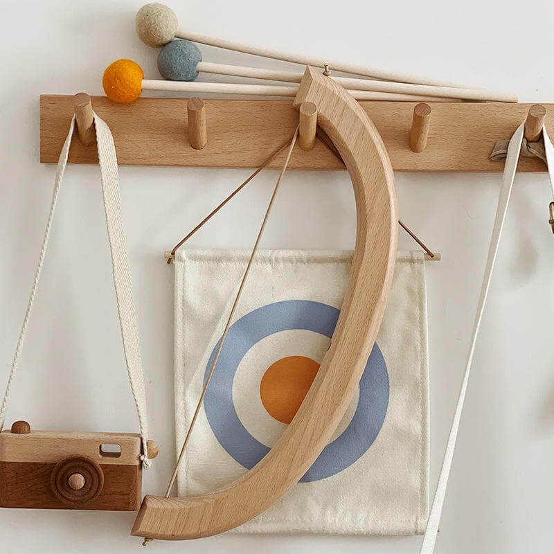 Wooden toys and accessories hang from a wooden peg rail against a white wall. Items include an InvenToy Montessori Bow and Arrow Set, a camera, Montessori toys like mallets with ball heads in various colors, and a canvas target with blue and orange circles. This eco-friendly wood setup has a minimalist and playful aesthetic that enhances hand-eye coordination.