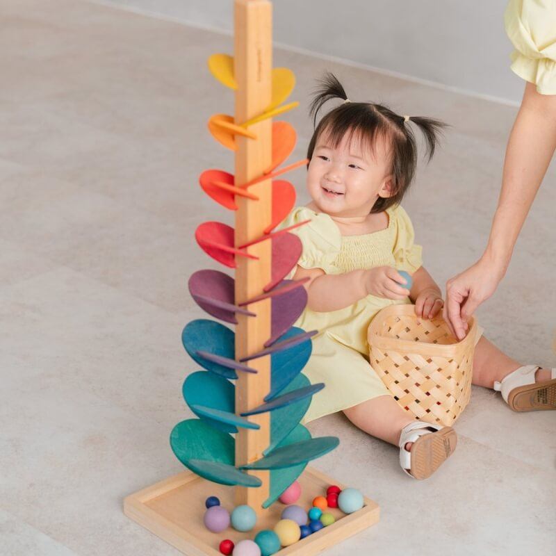 A toddler with pigtails in a yellow dress smiles while playing with the colorful InvenToy Montessori Rainbow Tree. An adult's hand, partially visible, assists with placing balls from a wicker basket into.
