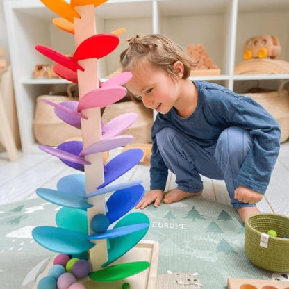 A toddler in a blue outfit is playing with a colorful InvenToy Montessori Rainbow Tree shaped like a tree in a warmly lit playroom. Various toys and baskets can be seen in the background.