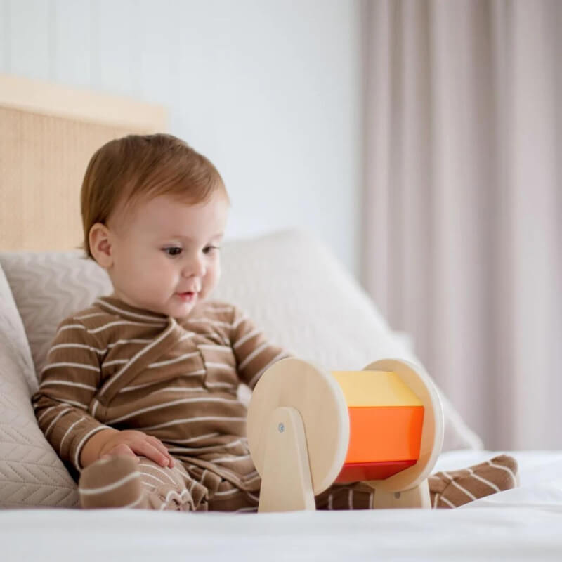 A toddler in a striped outfit sits on a bed, interacting with an InvenToy Montessori Spinning Drum. The child appears focused and curious. Neutral-colored bedding and a soft, blurred background create a serene setting.