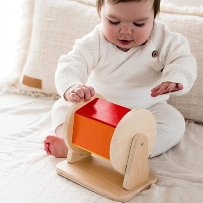 A baby in a white sweater plays with an InvenToy Montessori Spinning Drum on a soft rug, sitting upright and focused on hitting the drum.