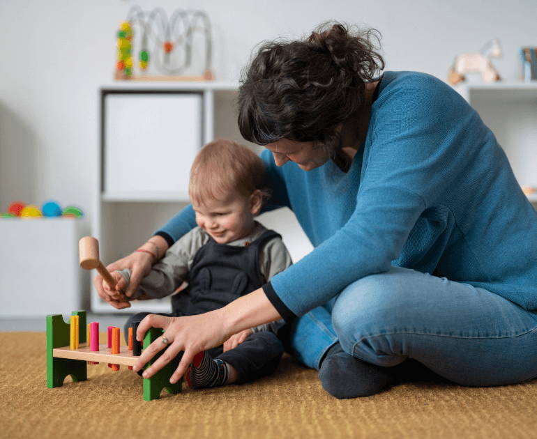 A woman in a blue sweater and jeans sits on the floor with a toddler, playing with a colorful wooden toy xylophone in a softly lit room.