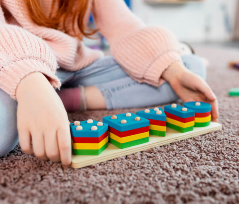 A young girl sits on a carpet, focusing on colorful building blocks arranged on a tactile board, primarily in green, red, and blue. her hands are poised to adjust a block.