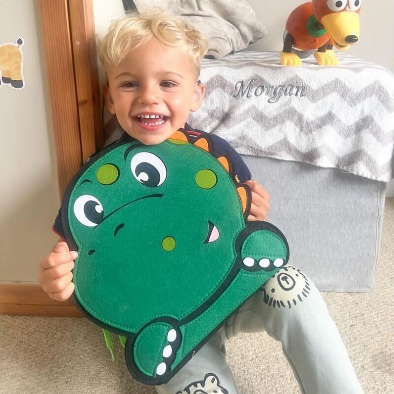 A cheerful young boy with blond hair smiling widely and holding a green frog-shaped backpack in front of him. A colorful InvenToy Montessori Dino Busy Board is visible on a bed in the background.