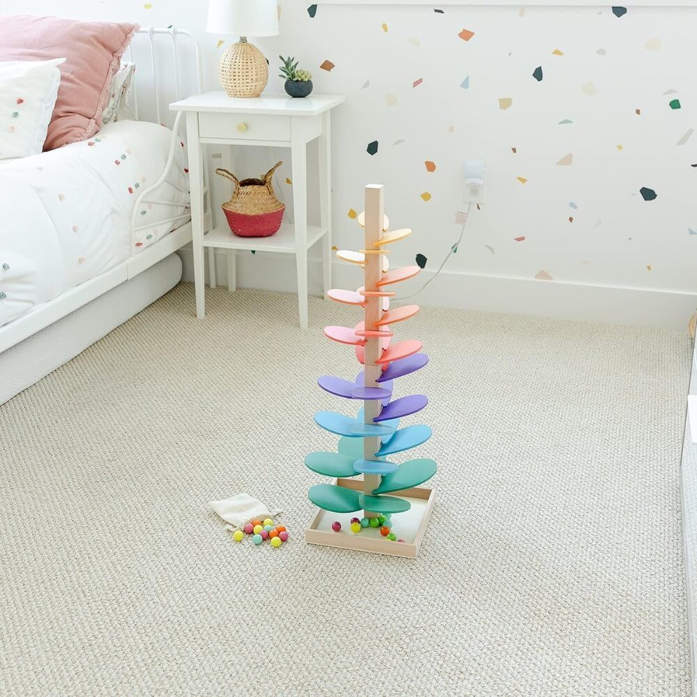 A colorful wooden InvenToy Montessori Rainbow Tree with multi-colored leaves stands on a carpeted floor in a child's bedroom, next to small balls and a book, with a decorated wall and furniture.