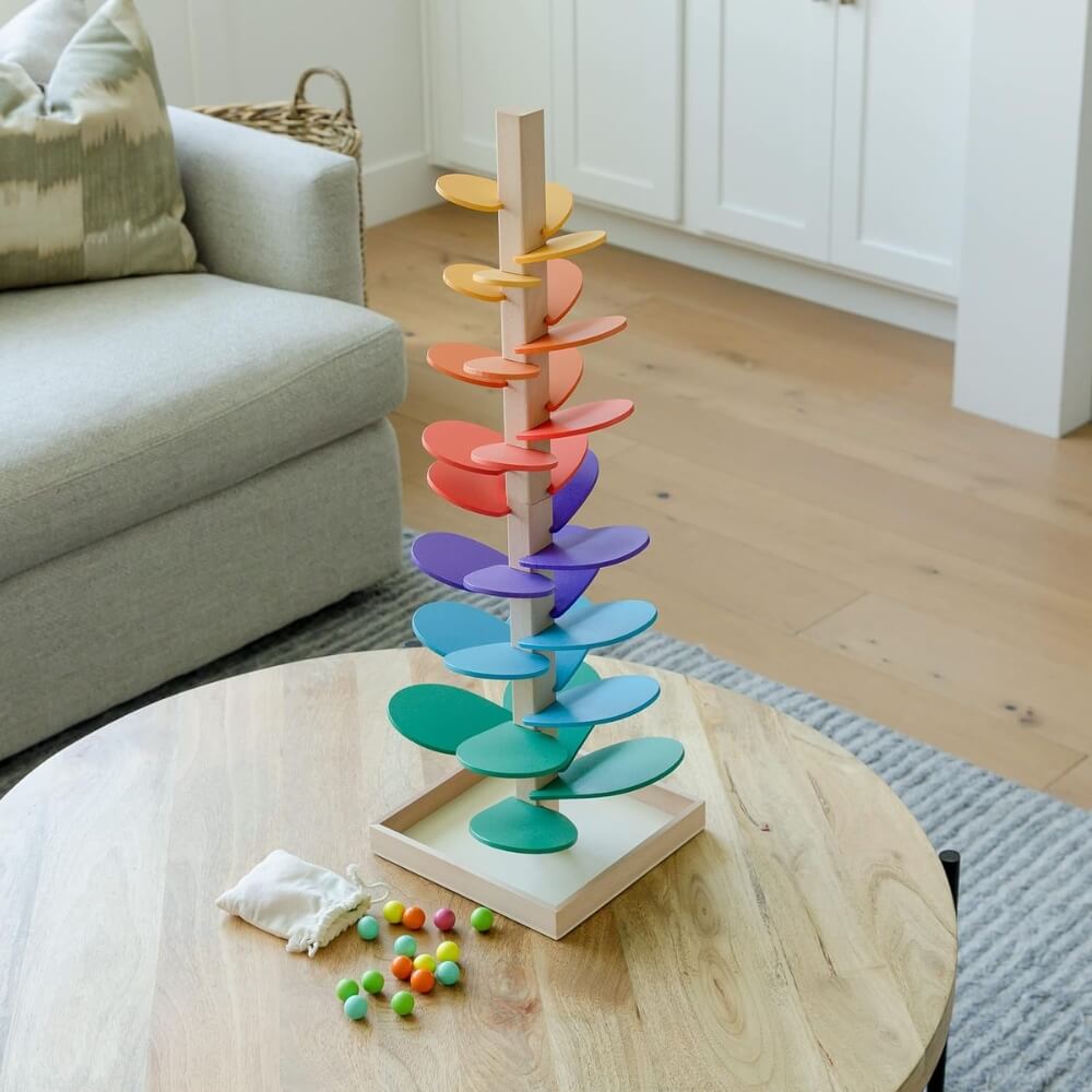 A colorful vertical InvenToy Montessori Rainbow Tree with spinning elements on a wooden coffee table in a cozy living room setting, accompanied by a cloth and scattered beads.
