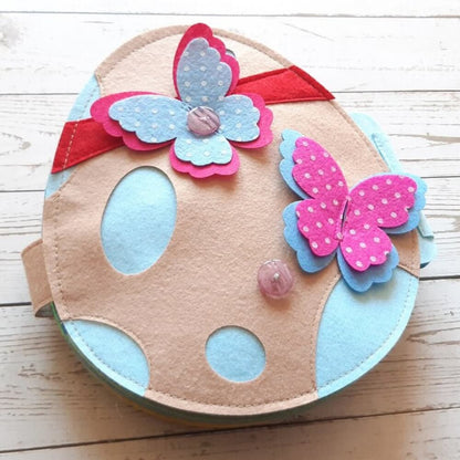 Handmade circular felt Easter egg decor with blue polka dots and colorful butterfly appliques on a white wooden background, perfect as InvenToy's Montessori Rabbit's Book.