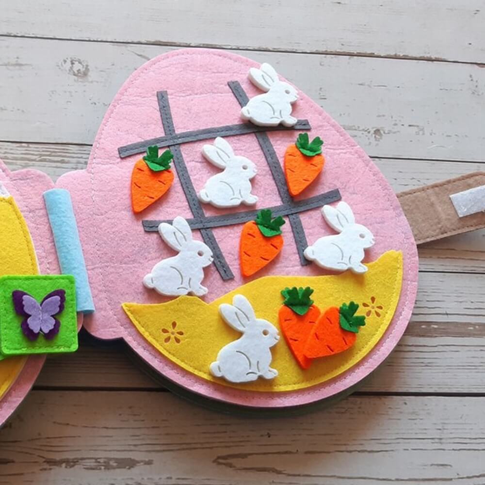 A colorful felt Easter egg featuring a tic-tac-toe game with white bunny and orange carrot pieces on a pink and yellow background, displayed on a wooden surface. This InvenToy Montessori Rabbit's Book enhances.