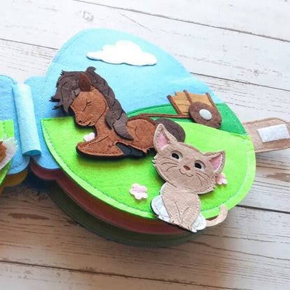 A colorful felt book page featuring a blue and green landscape with a brown felt horse lying down, and a beige kitten sitting nearby, both amidst small pink flowers. This inventive Montessori Rabbit's Book encourages sensory.