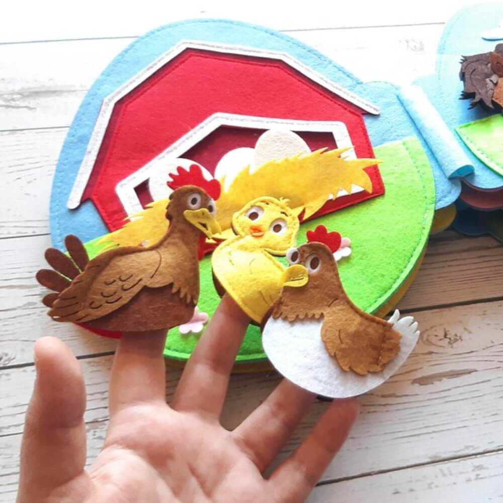 A hand holding colorful felt crafts featuring two brown birds and a yellow chick against a backdrop of a red barn and green fields, perfect as InvenToy's Montessori Rabbit's Book baby toys.