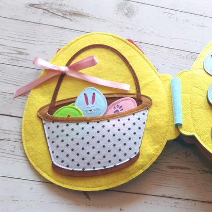 A colorful felt Easter basket with a blue bunny and two eggs on a yellow circular background, decorated with a pink ribbon on a wooden surface, featuring InvenToy's Montessori Rabbit's Book.