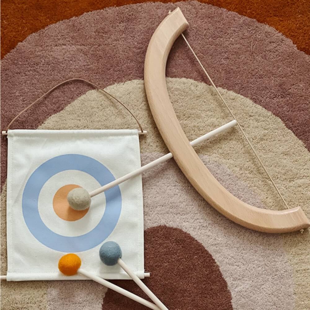 An InvenToy Montessori Bow and Arrow Set, crafted from eco-friendly wood, lies on a multicolored circular-patterned rug. The set includes a wooden bow with a string and two toy arrows with felt tips. A fabric target with concentric circles in shades of blue and orange is also part of this Montessori toy designed for enhancing hand-eye coordination.