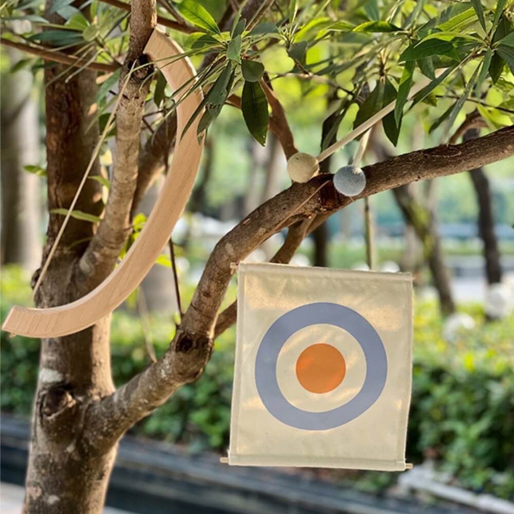 A tree has two items hanging from its branches: a wooden crescent on a rope and an InvenToy Montessori Bow and Arrow Set, featuring a fabric square with a blue and orange target design, perfect for improving hand-eye coordination. The background shows a blurred view of more trees and a walkway.