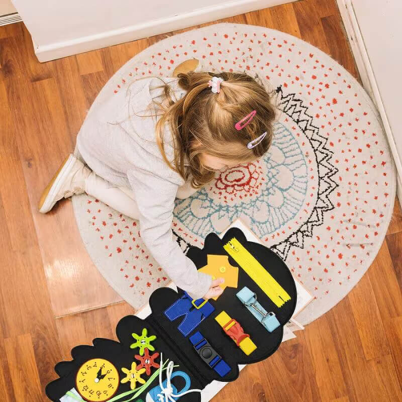 A young child with blonde hair is playing with a colorful, InvenToy Montessori Dino Busy Board on a round, patterned rug. The toy features various shapes and clocks, aimed at learning skills.