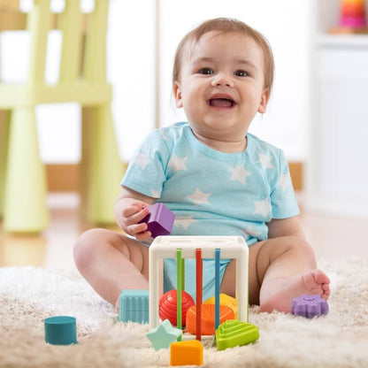A joyful baby in a blue star-patterned outfit sitting on a carpet, playing with colorful InvenToy Montessori Shape Blocks.