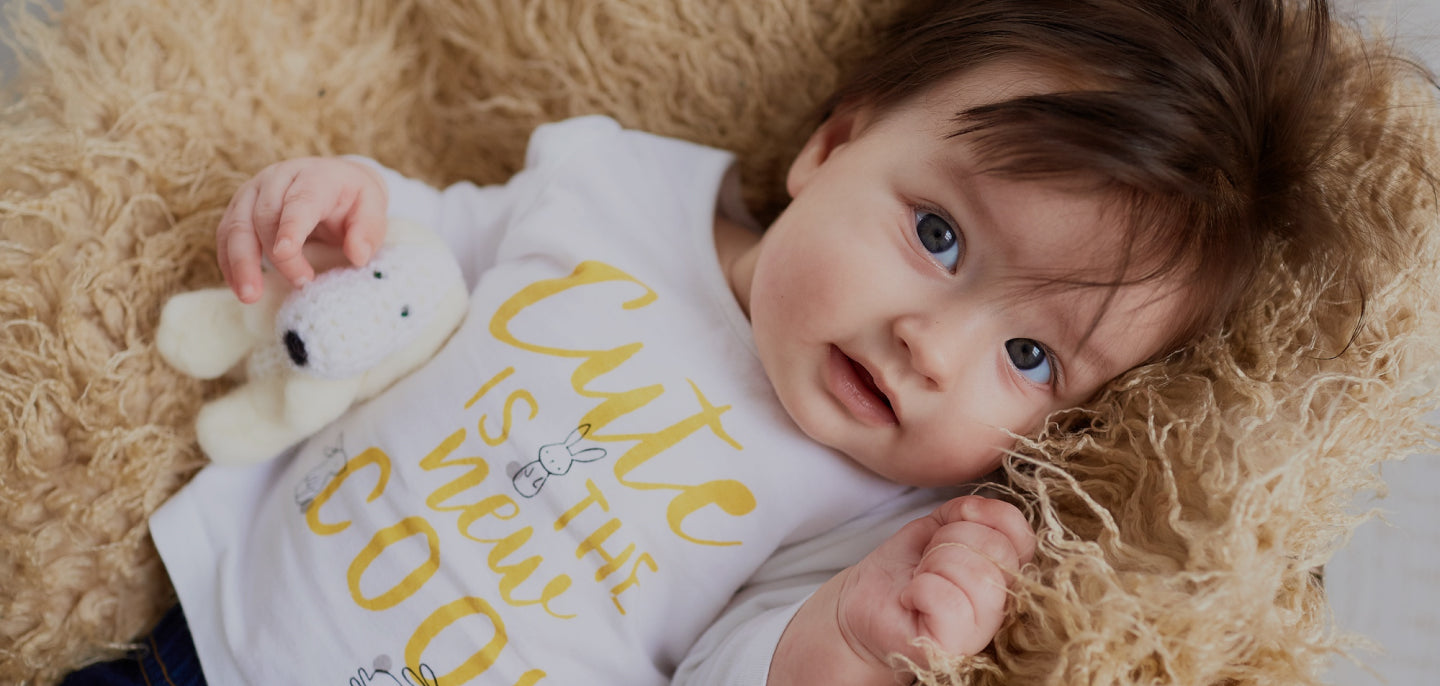 A baby with thick brown hair lying on a soft, beige blanket, holding a small white stuffed animal. the baby wears a white onesie with the text "cute is the new cool.