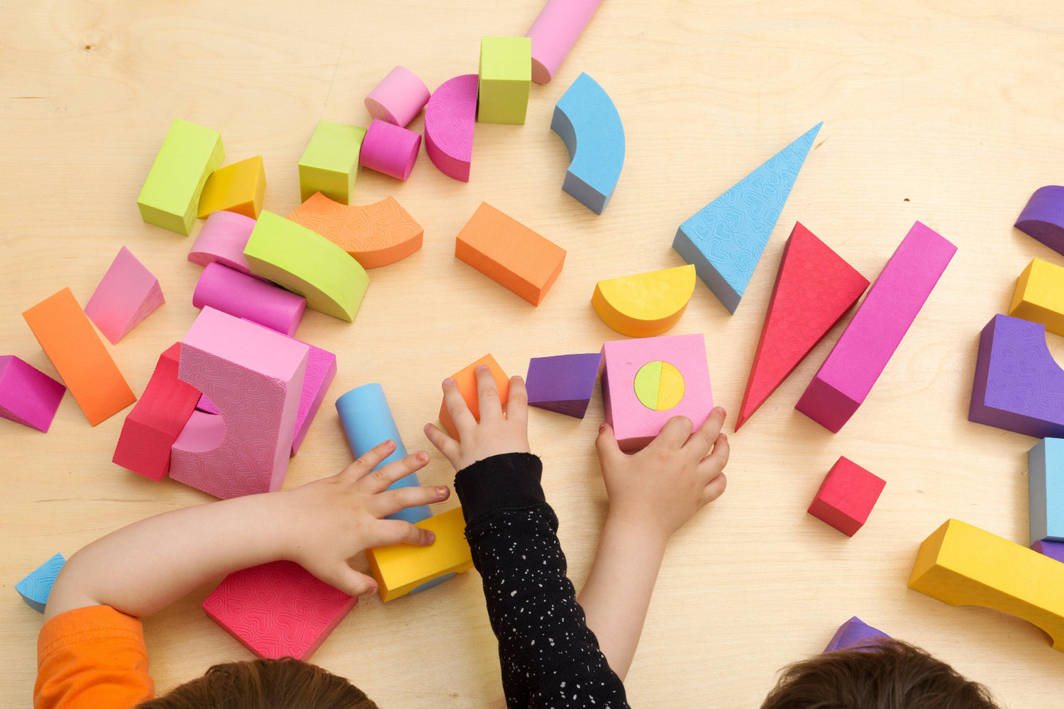 A top view of two children's hands playing with colorful wooden blocks scattered on a wooden floor, building and arranging them creatively.