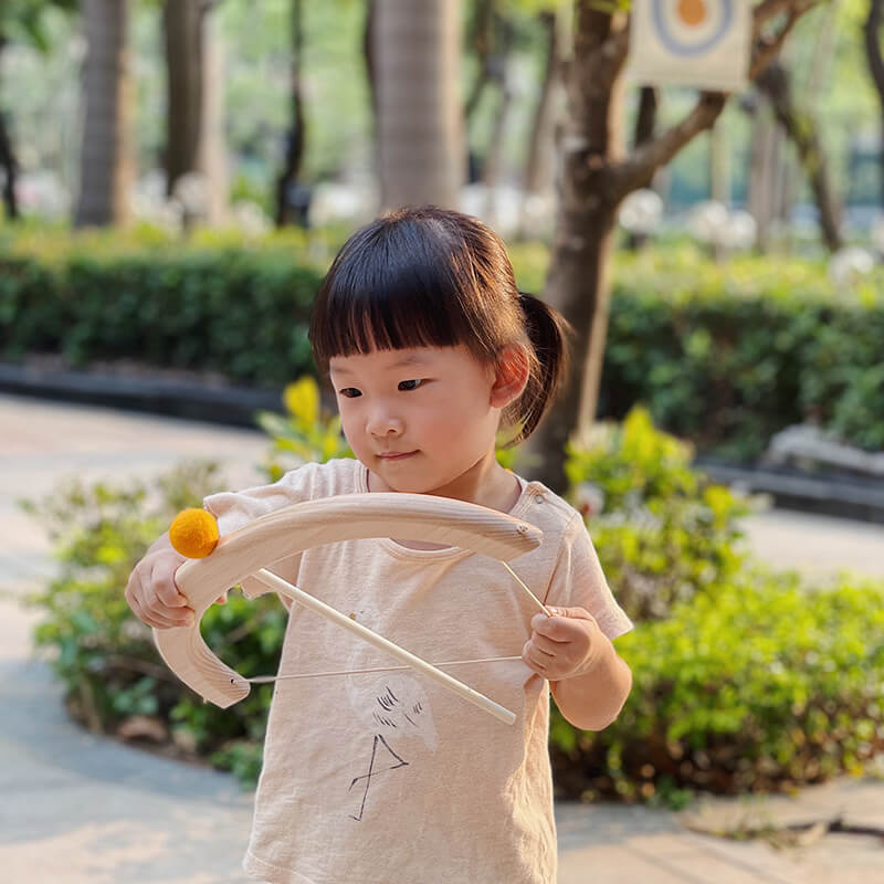 A young child with short hair is concentrating on using the InvenToy Montessori Bow and Arrow Set outdoors. Standing on a paved path with trees and greenery in the background, the child develops hand-eye coordination. The eco-friendly wood toy is bathed in sunlight, creating a warm, serene atmosphere.