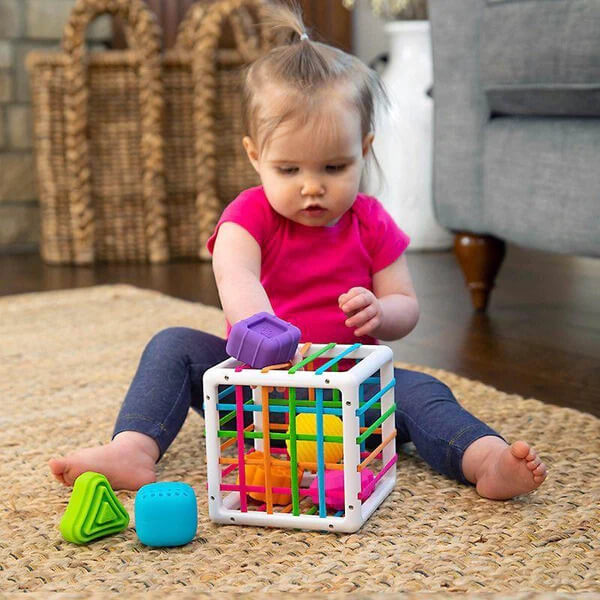A baby sits on a textured rug, playing with a colorful InvenToy Montessori Shape Blocks, focused on fitting a purple block into the toy.